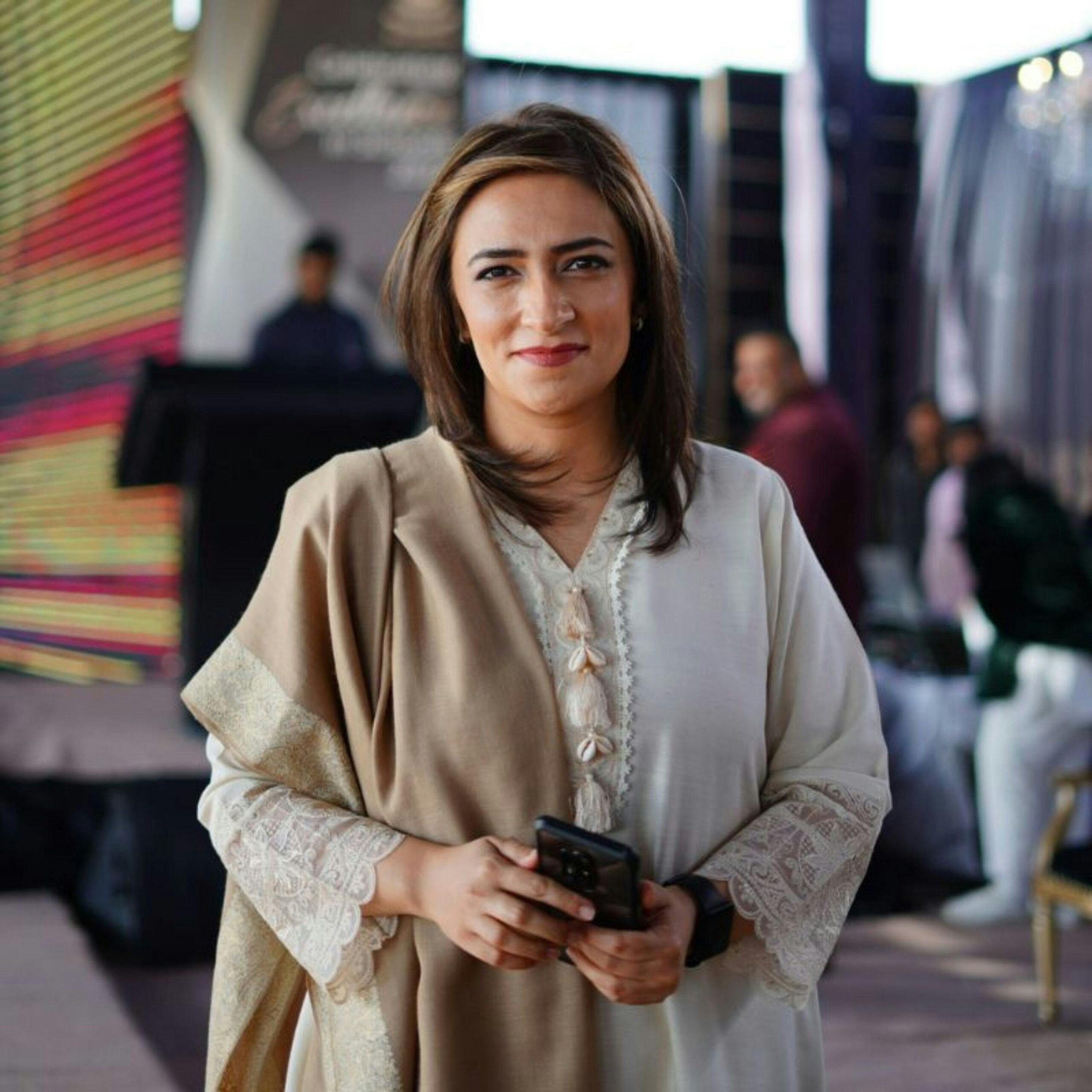 Elegant businesswoman holding smartphone, smiling at an event in Dubai. Professional attire and warm ambiance.